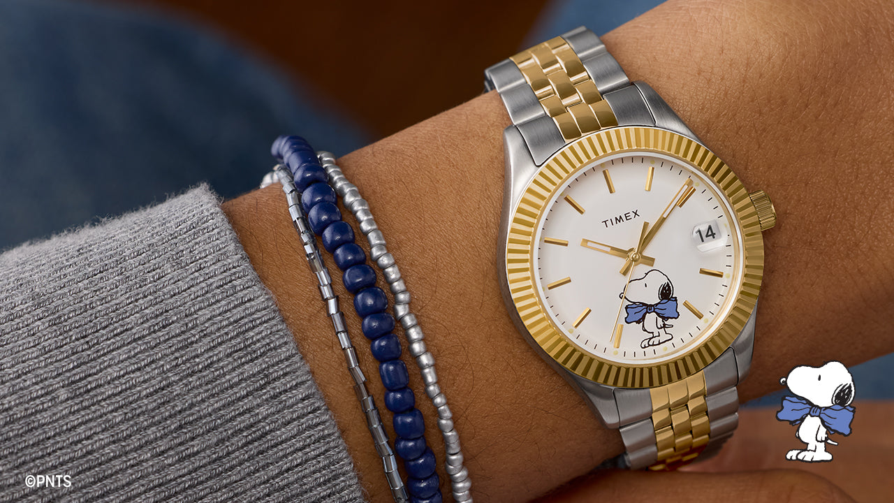 Timex x Peanuts® Timex Legacy watch on wrist with silver and gold-tone bracelet, gold fluted bezel, white dial featuring Snoopy wearing a blue bow tie at 6 o’clock, date window at 3 o’clock, paired with silver and navy beaded bracelets.