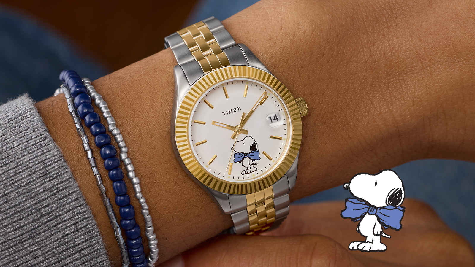 Timex x Peanuts® Timex Legacy watch on wrist with silver and gold-tone bracelet, gold fluted bezel, white dial featuring Snoopy wearing a blue bow tie at 6 o’clock, date window at 3 o’clock, paired with silver and navy beaded bracelets.