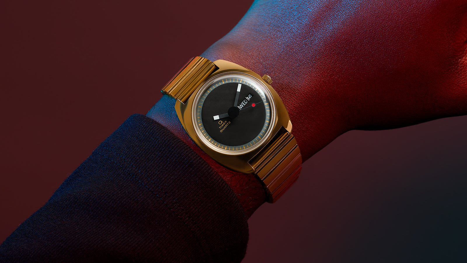 Q Timex® 1975 Enigma watch on wrist, showing the sleek gold-tone case and bracelet, black dial with minimalist indices, geometric hands, small red dot at 6 o’clock, and day-date window at 3 o’clock, against a deep red and blue gradient backdrop.