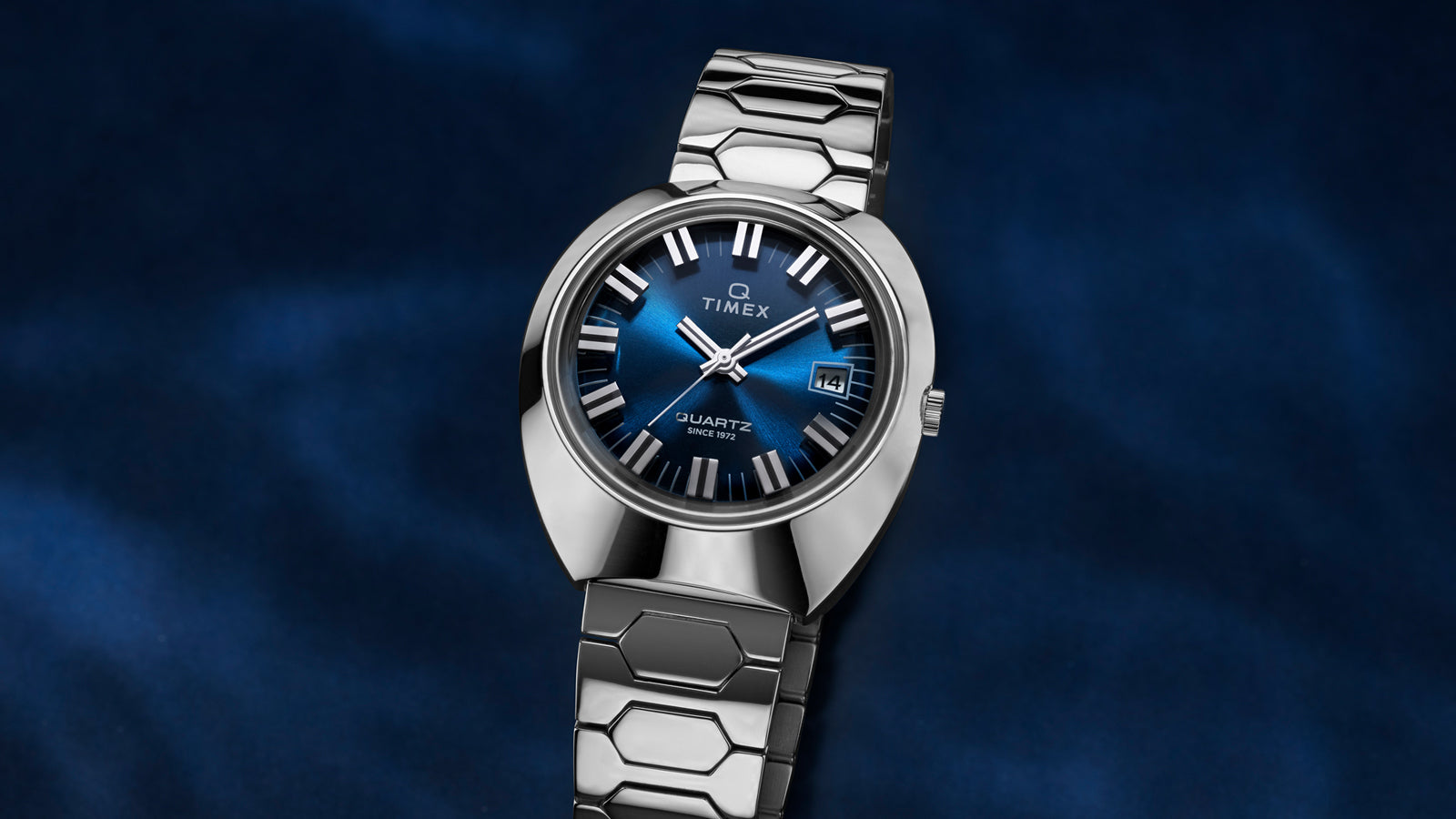 Q Timex® 1972 Time Machine Reissue wristwatch with a polished stainless-steel case, bold faceted design, and hexagonal-link stainless-steel bracelet. Features a deep blue sunray dial, silver-tone hour markers with black accents, luminous hands, and a date window at 3 o’clock, displayed against a dark blue gradient background.