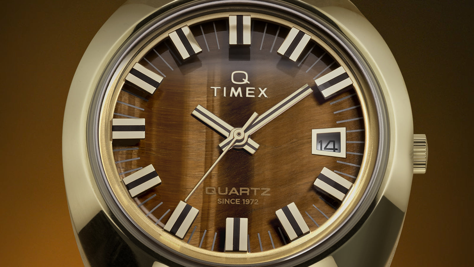 Close-up of the Q Timex® 1972 Time Machine Reissue gold-tone watch dial showing the rich tiger’s-eye effect, bold silver-tone hour markers with black accents, luminous hands, and a date window at 3 o’clock, with “Q Timex” and “Quartz Since 1972” printed on the dial.