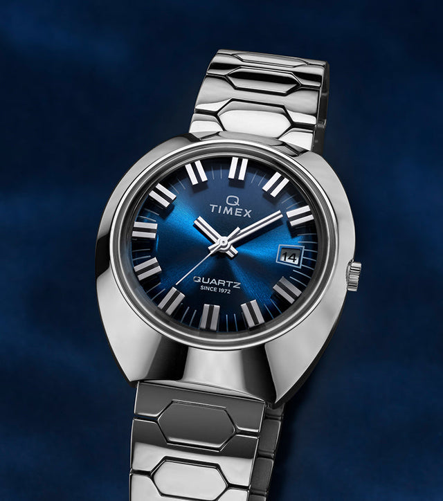 Q Timex® 1972 Time Machine Reissue wristwatch with a polished stainless-steel case, bold faceted design, and hexagonal-link stainless-steel bracelet. Features a deep blue sunray dial, silver-tone hour markers with black accents, luminous hands, and a date window at 3 o’clock, displayed against a dark blue gradient background.