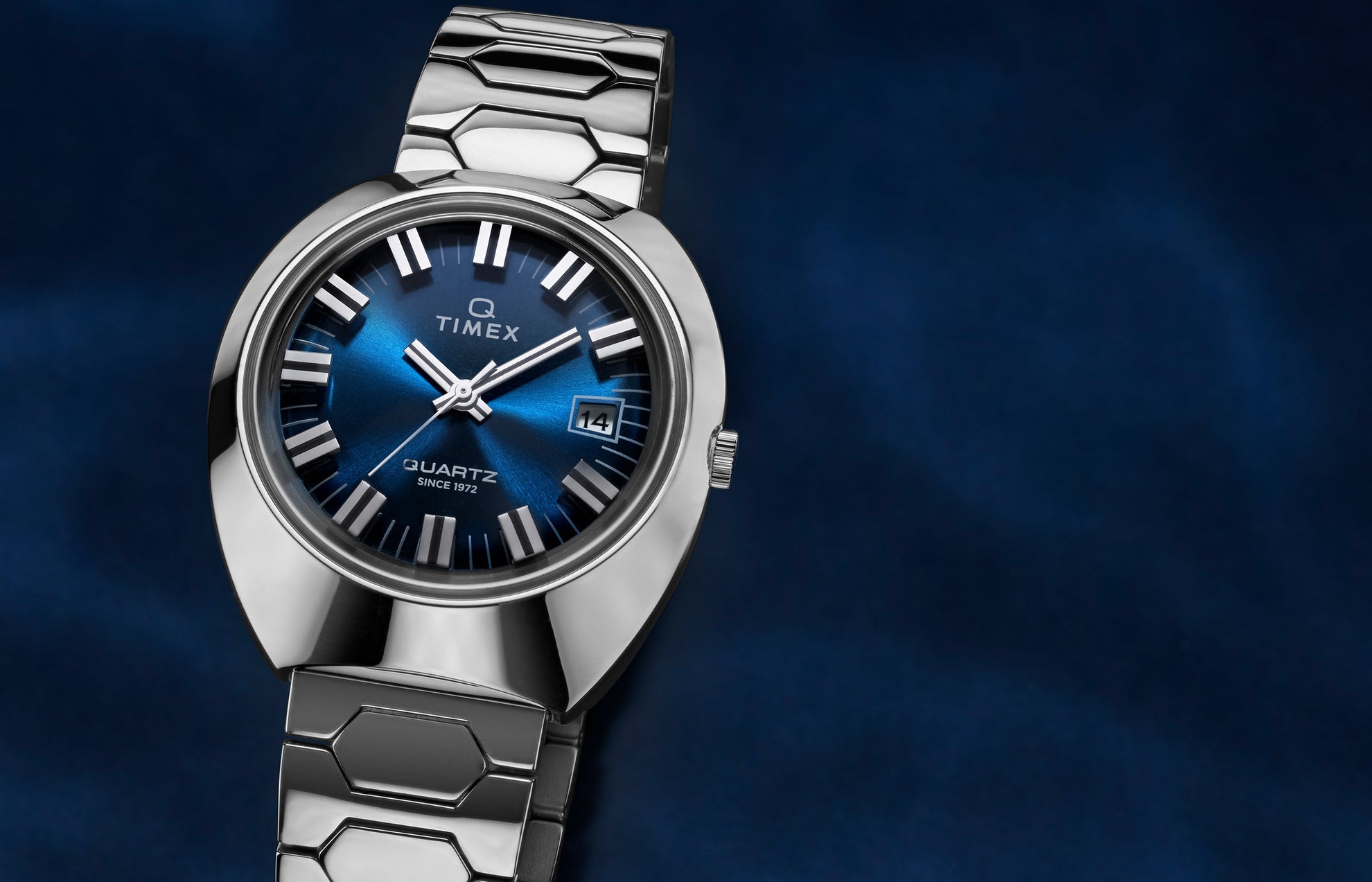 Q Timex® 1972 Time Machine Reissue wristwatch with a polished stainless-steel case, bold faceted design, and hexagonal-link stainless-steel bracelet. Features a deep blue sunray dial, silver-tone hour markers with black accents, luminous hands, and a date window at 3 o’clock, displayed against a dark blue gradient background.