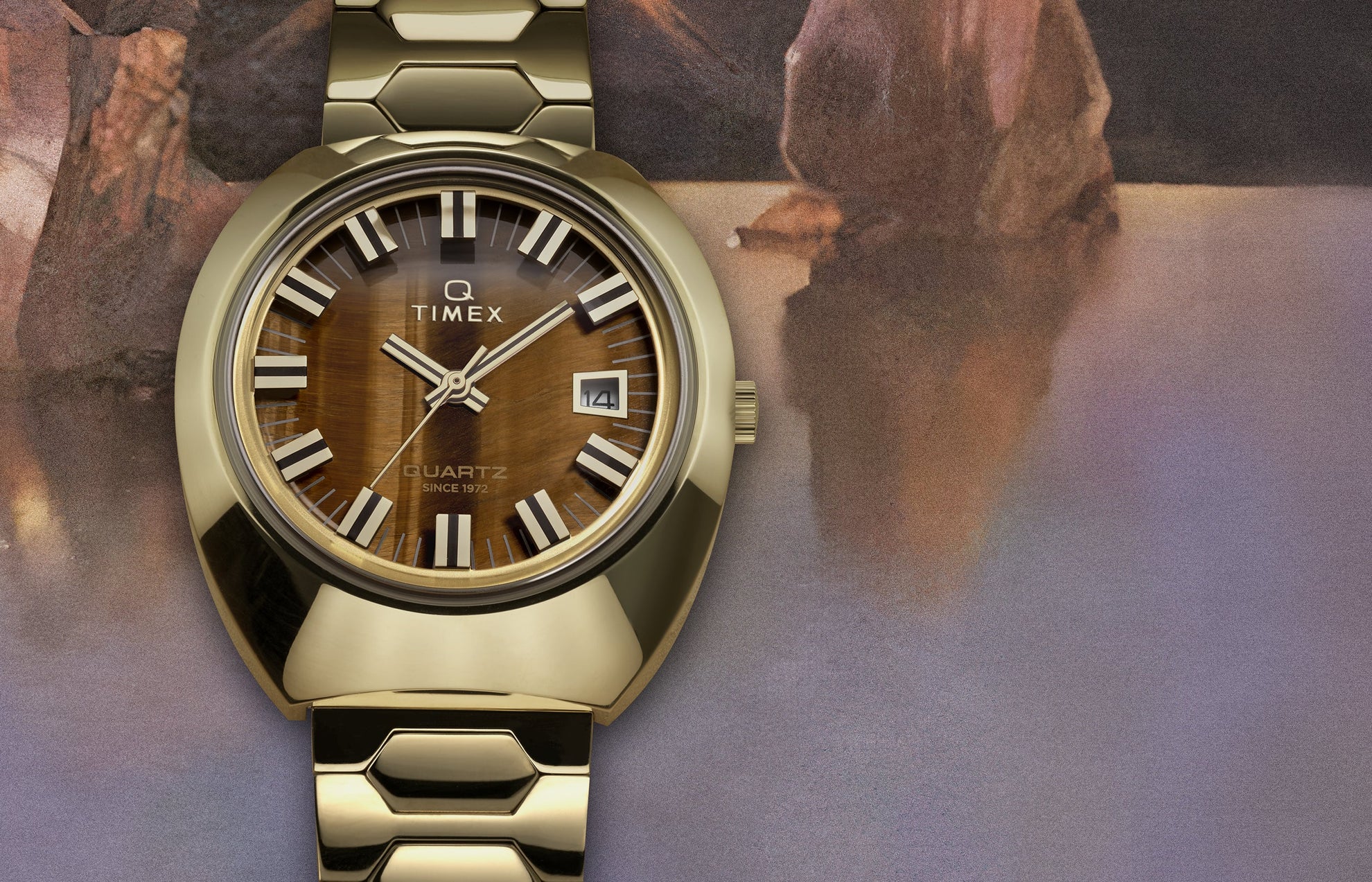 Q Timex® 1972 Time Machine Reissue wristwatch in polished gold-tone stainless steel with a faceted case and hexagonal-link bracelet. Features a tiger’s-eye style brown dial with silver-tone hour markers accented in black, luminous hands, and a framed date window at 3 o’clock, displayed against a textured, artistic background.