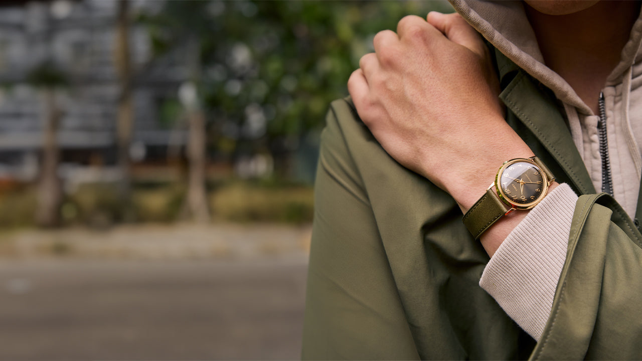 A close-up of a person wearing the Timex x Todd Snyder watch, featuring a green leather strap and gold-tone case, layered over a green jacket and beige hoodie in an outdoor urban setting.