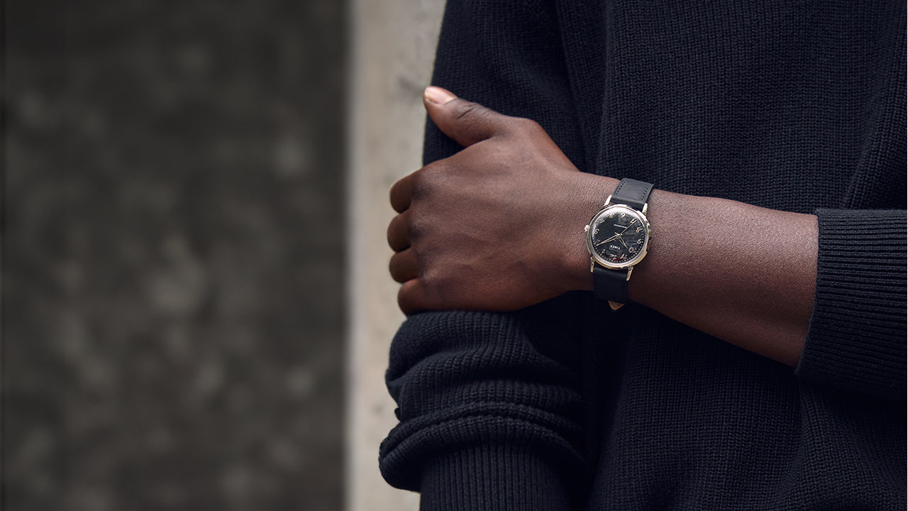 Close-up lifestyle image of a person wearing the Timex x Todd Snyder watch with a black dial and black leather strap, styled with a black textured sweater for a modern monochrome look.