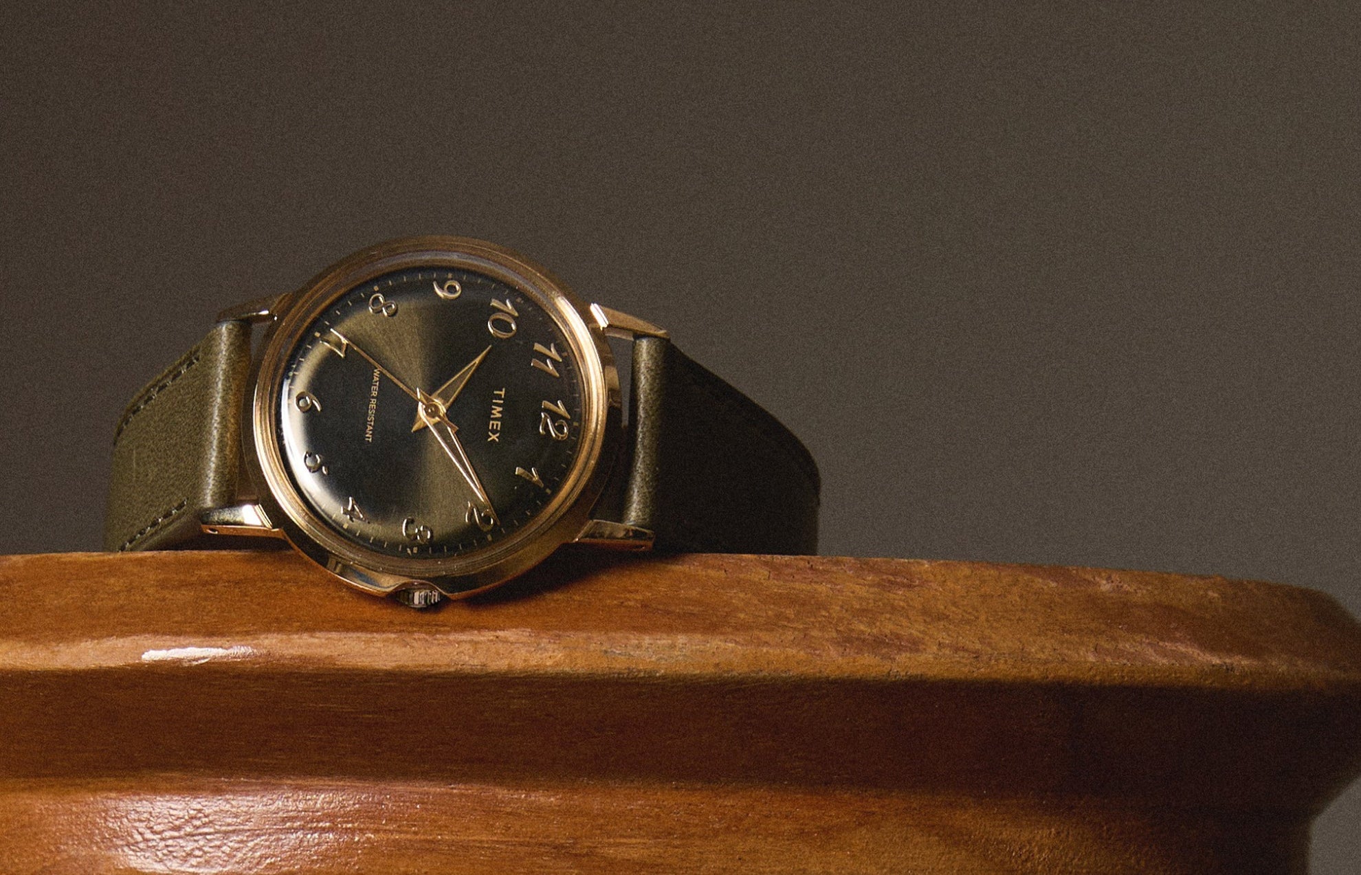 Timex x Todd Snyder watch resting on a wooden surface, showcasing the green dial, gold-tone case, and olive leather strap with a sophisticated, vintage-inspired look.