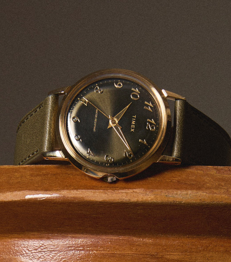 Timex x Todd Snyder watch resting on a wooden surface, showcasing the green dial, gold-tone case, and olive leather strap with a sophisticated, vintage-inspired look.
