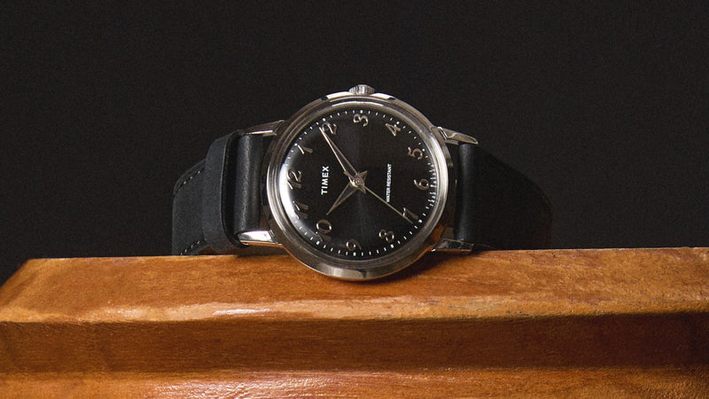 Timex x Todd Snyder watch resting on a wooden surface, highlighting the polished silver-tone case, black sunray dial, and classic Arabic numerals with a sleek black leather strap.