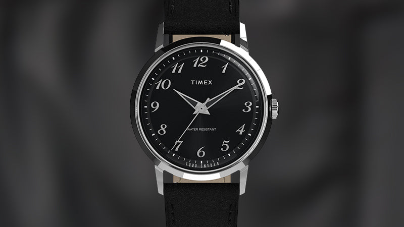 Product shot of the Timex x Todd Snyder watch against a blurred dark background, showcasing its crisp black dial, silver-tone case, and black strap in sharp detail.