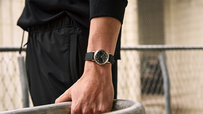 Urban lifestyle photo of a person leaning on a metal railing, wearing the Timex x Todd Snyder watch with black strap and dial, styled with all-black athleisure for a contemporary streetwear vibe.