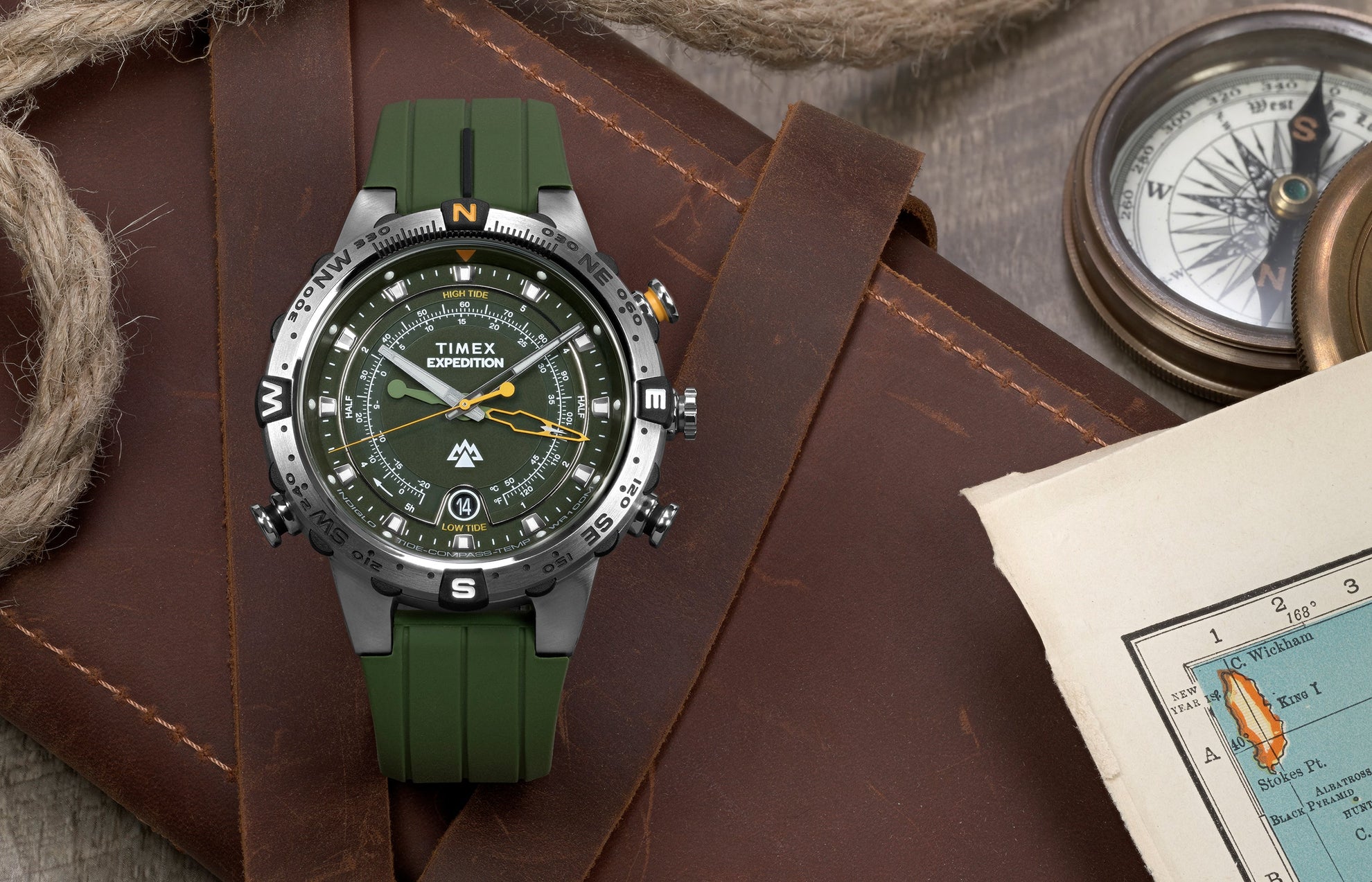 Timex Expedition® Tide-Temp-Compass watch with a green strap placed on a leather surface beside a vintage compass, nautical rope, and a folded map.