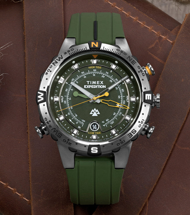 Timex Expedition® Tide-Temp-Compass watch with a green strap placed on a leather surface beside a vintage compass, nautical rope, and a folded map.