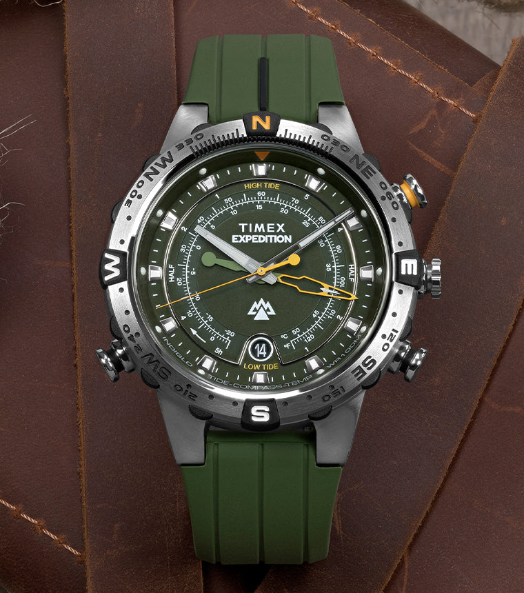 Timex Expedition® Tide-Temp-Compass watch with a green strap placed on a leather surface beside a vintage compass, nautical rope, and a folded map.