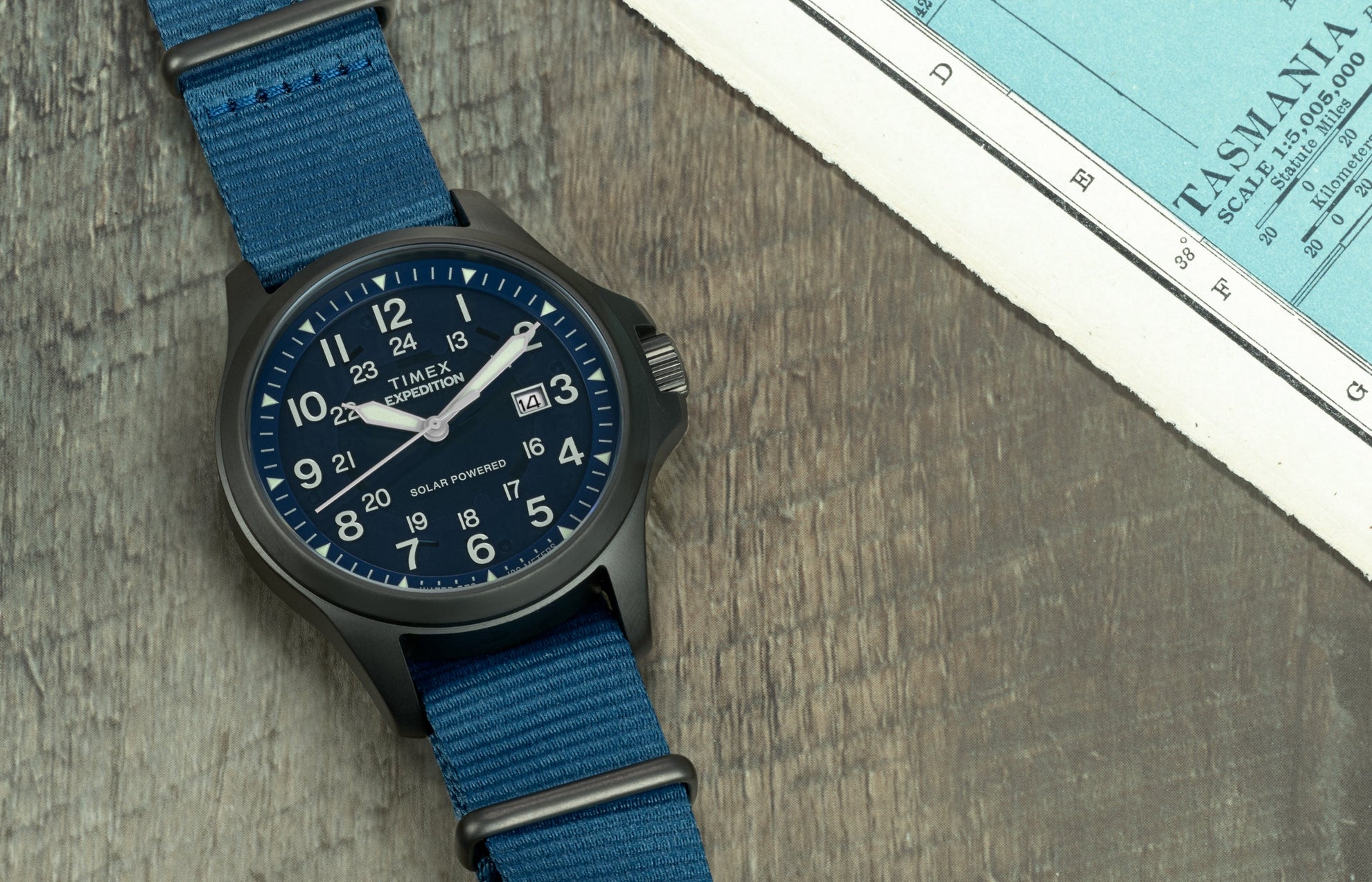Expedition Field Post Solar watch with blue dial and strap on map background – Timex Expedition® Field Post Solar watch featuring a deep blue dial, white numerals, and fabric strap, placed on a wooden surface next to a vintage map.