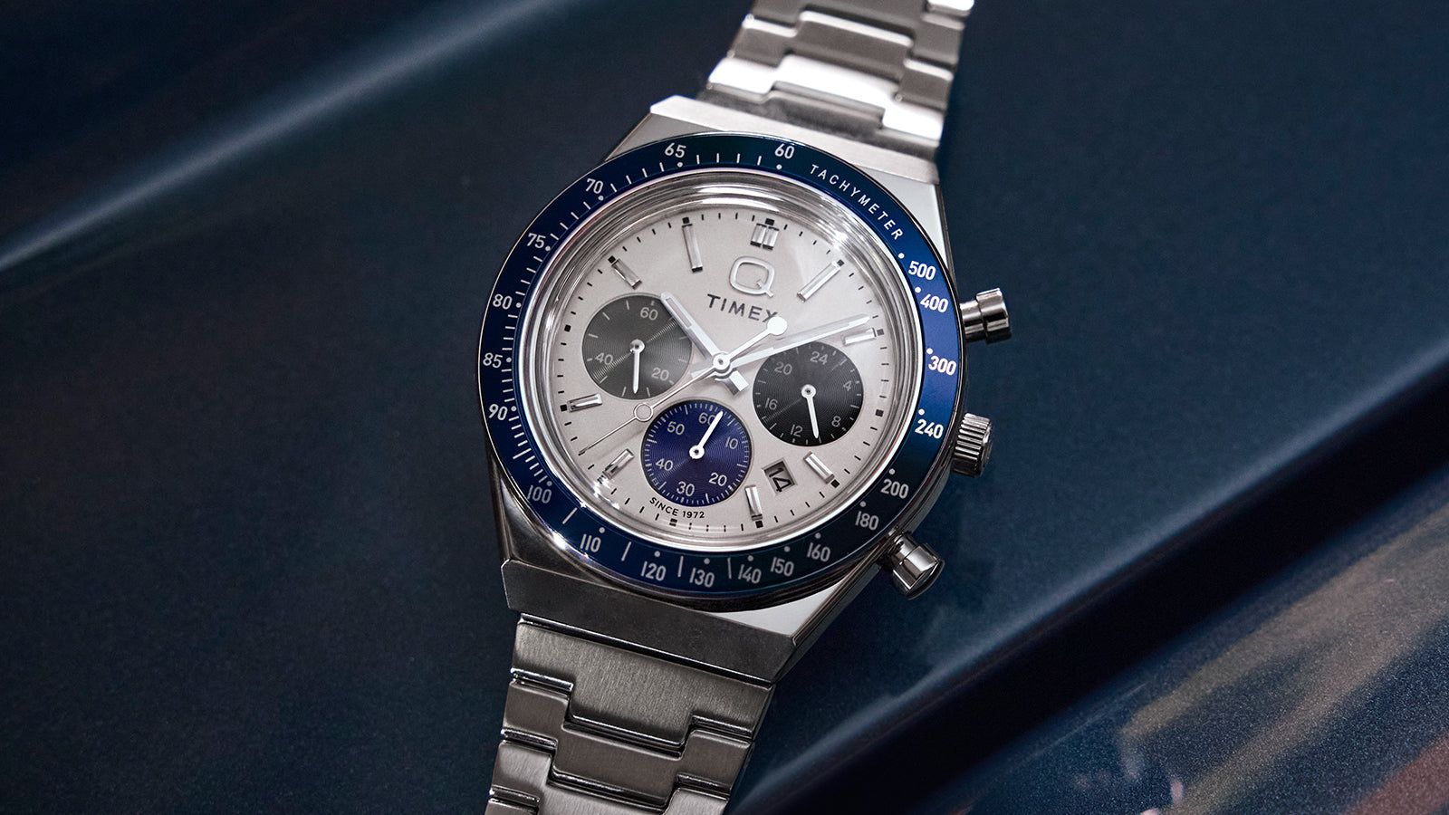 Q Timex® Chronograph on a metallic blue surface—white dial with three sub-dials, navy tachymeter bezel, date at 4 o’clock, stainless-steel bracelet.