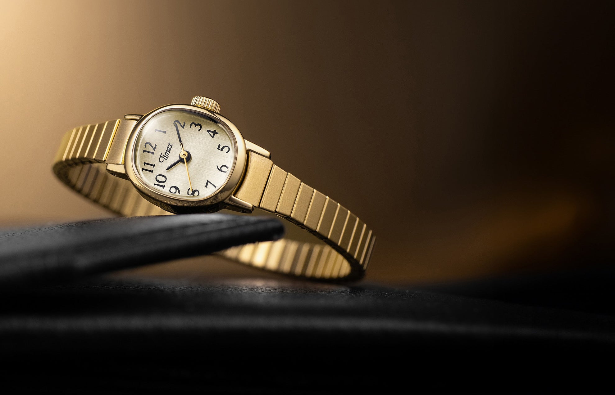 Timex Cavatina Mini gold-tone watch with expansion bracelet, resting on a black leather surface with a warm golden background.