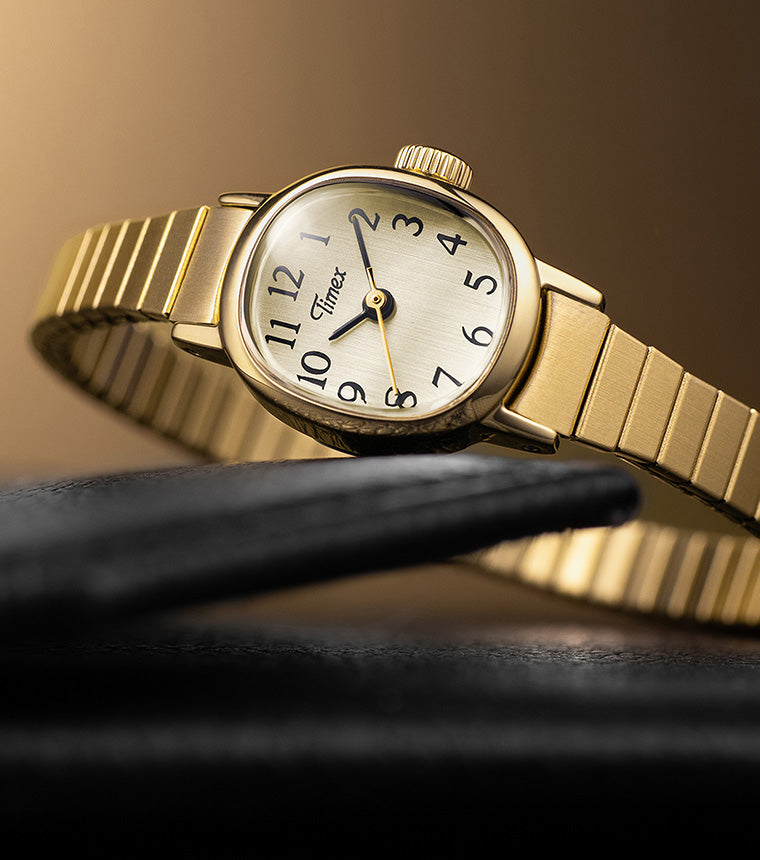 Timex Cavatina Mini gold-tone watch with expansion bracelet, resting on a black leather surface with a warm golden background.