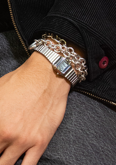 A person sitting casually with hands resting on their lap, wearing the Teeny Tiny Timex digital watch paired with a chunky silver chain bracelet. The look blends nostalgic digital style with contemporary streetwear, highlighting Dimepiece’s urban-chic influence.
