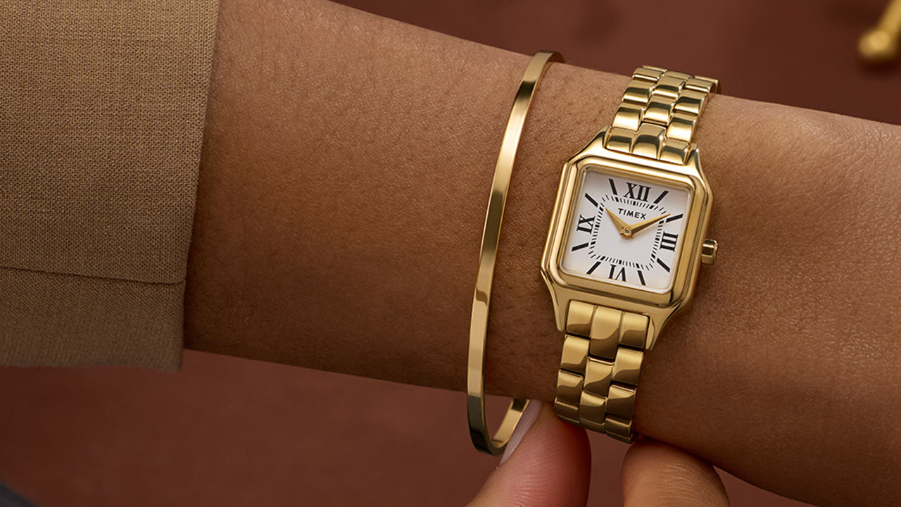 Timex Legacy Addison gold-tone rectangular watch with white Roman numeral dial, worn on the wrist beside a slim gold bangle.
