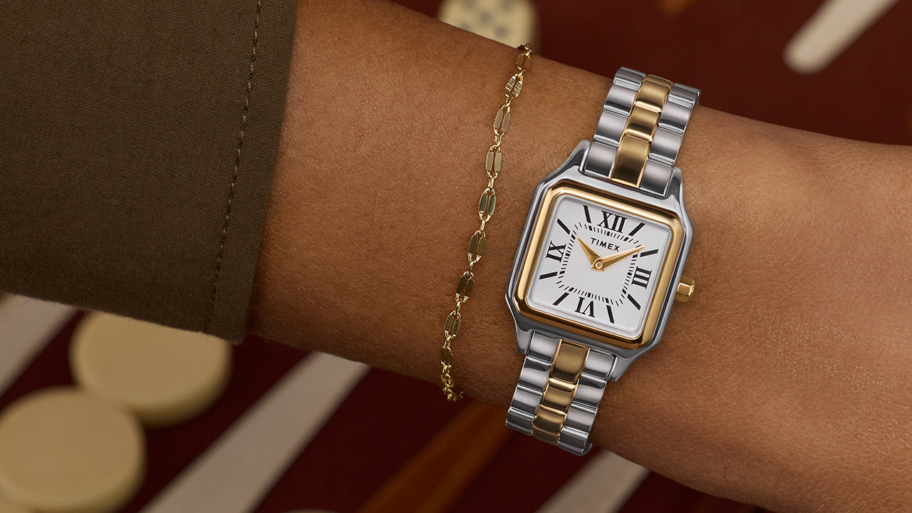 Timex Legacy Addison two-tone square watch with white Roman-numeral dial, worn on the wrist beside a delicate gold chain bracelet.