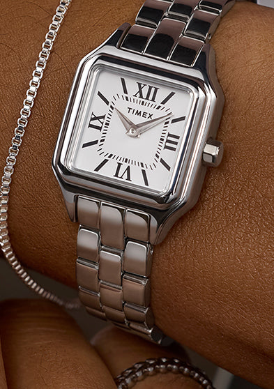 Timex Legacy Addison silver-tone square watch with white Roman-numeral dial, worn on the wrist beside a slim chain bracelet.