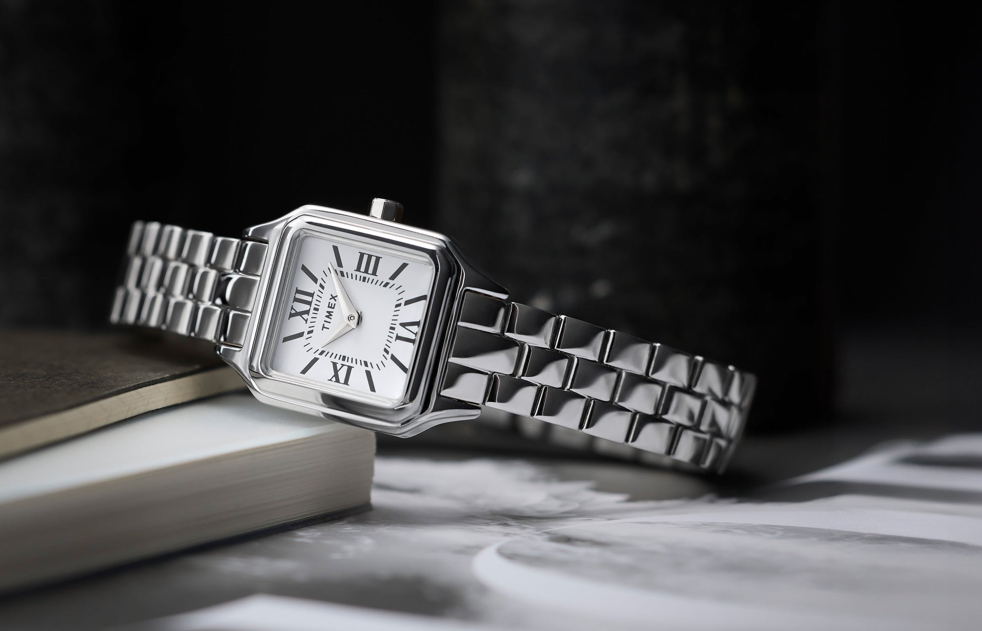 Timex Legacy Addison silver-tone women’s watch with square, rounded-corner case, white dial with black Roman numerals, and polished link bracelet, styled on books.