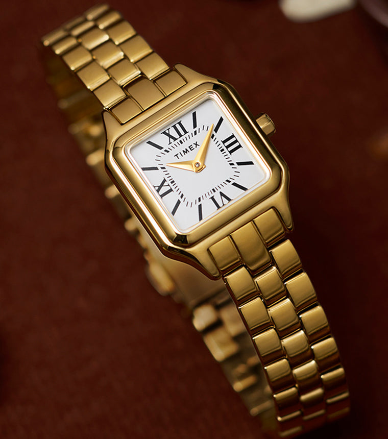 Close-up of the Timex Legacy Addison gold-tone watch showing square case, white dial with black Roman numerals, and gold hands on a link bracelet.