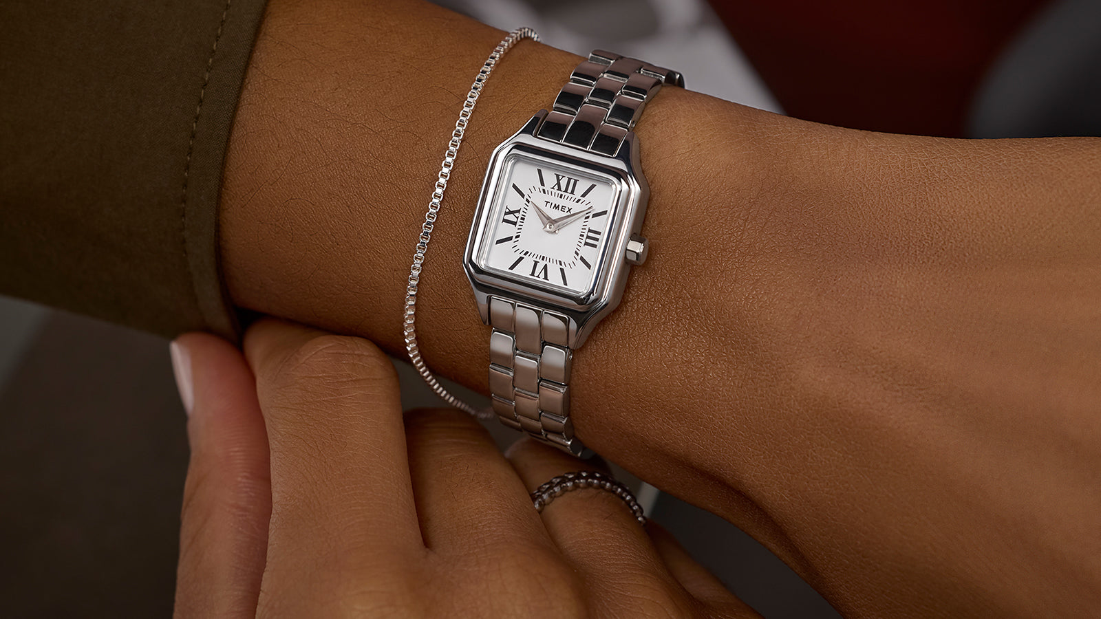 Timex Legacy Addison silver-tone square watch with white Roman-numeral dial, worn on the wrist beside a slim chain bracelet.