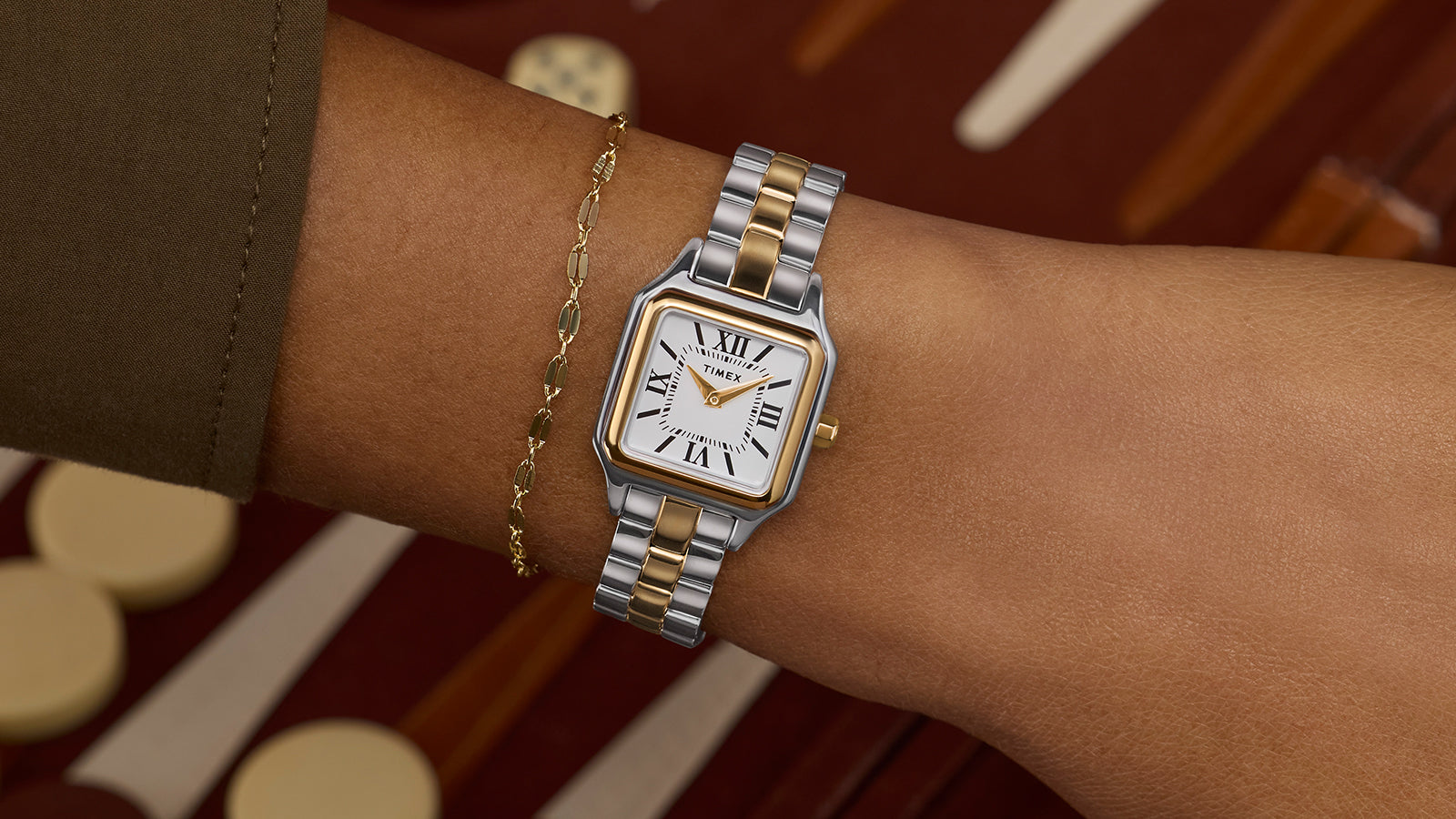 Timex Legacy Addison two-tone square watch with white Roman-numeral dial, worn on the wrist beside a delicate gold chain bracelet.