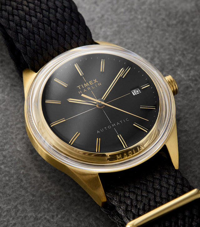 Timex Marlin® Jet Automatic watch with a black dial, gold-tone case, gold hands and indices, and black woven strap, displayed on a textured dark background.