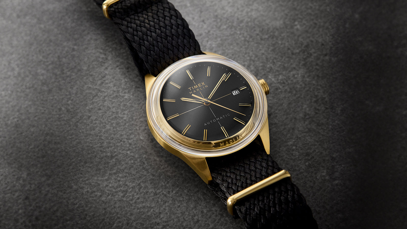 Timex Marlin® Jet Automatic watch with a black dial, gold-tone case, gold hands and indices, and black woven strap, displayed on a textured dark background.