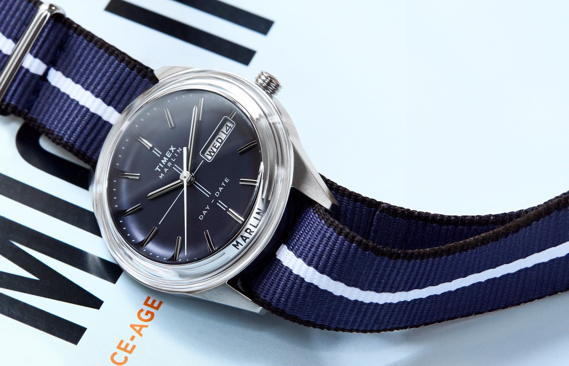 Timex Marlin® Jet Quartz watch with a deep blue dial, silver-tone markers, and day-date window, paired with a navy blue fabric strap featuring a white center stripe, displayed on a magazine cover.