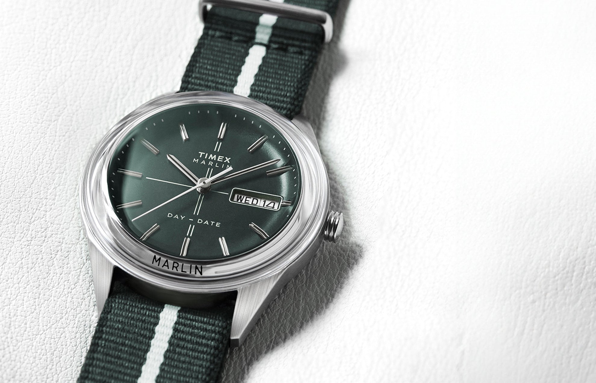 Timex Marlin® Jet Quartz watch with a deep green dial, silver-tone markers, and day-date window, paired with a dark green fabric strap featuring a white center stripe, displayed on a light leather surface.