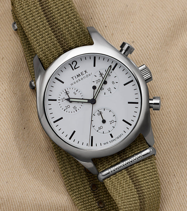 Timex Weekender New England Chronograph with white dial and three sub-dials on an olive fabric strap, styled on tan canvas.
