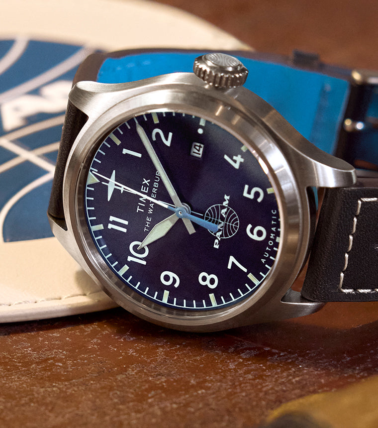 Close-up of the Timex x Pan Am® Waterbury Ace with a navy blue dial, bold white numerals, stainless steel case, and black leather strap featuring white stitching and embossed airplane detail.