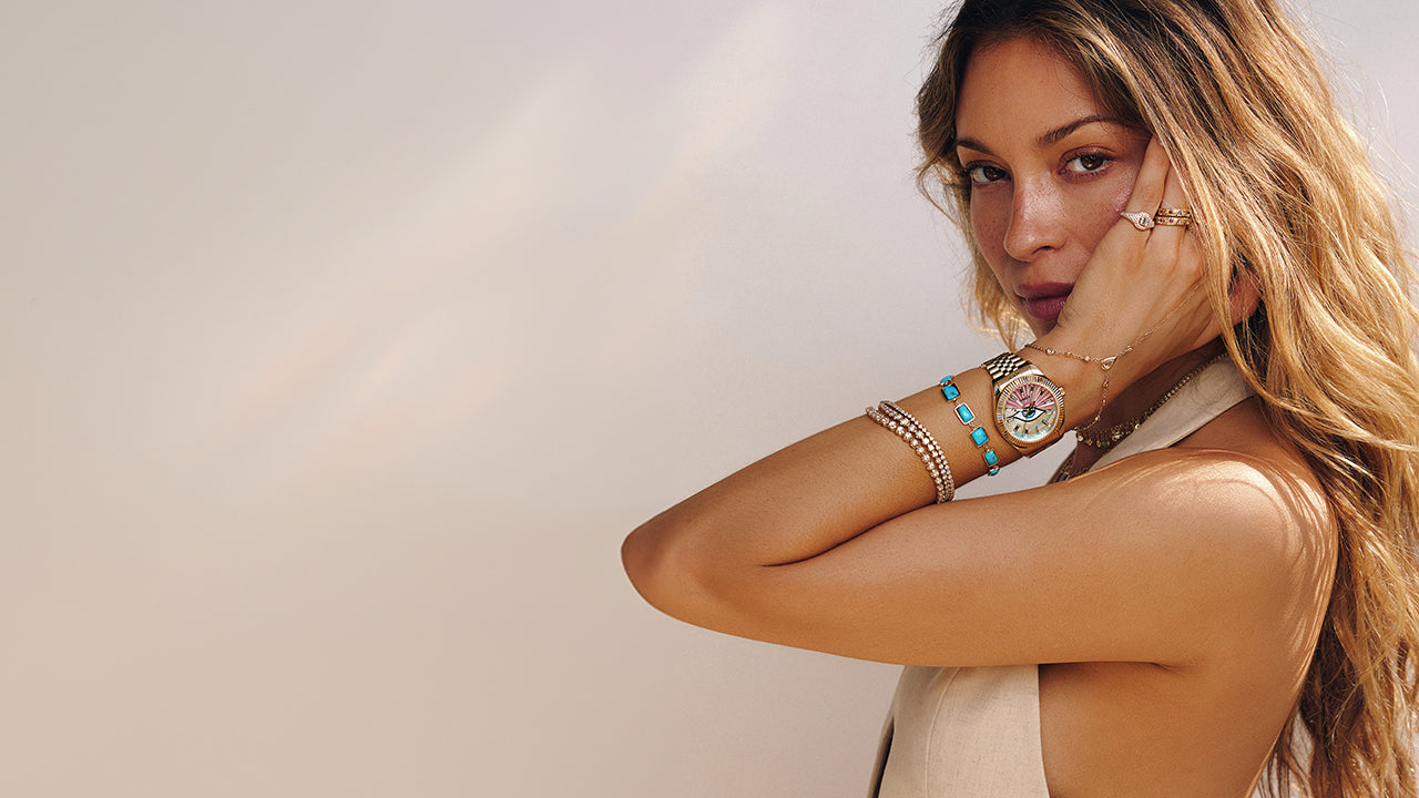 Woman wearing the Timex x Jacquie Aiche gold watch with a pink and multicolored mother-of-pearl dial featuring an eye motif, styled with layered gold and turquoise bracelets, posing with her hand near her face against a soft neutral background.
