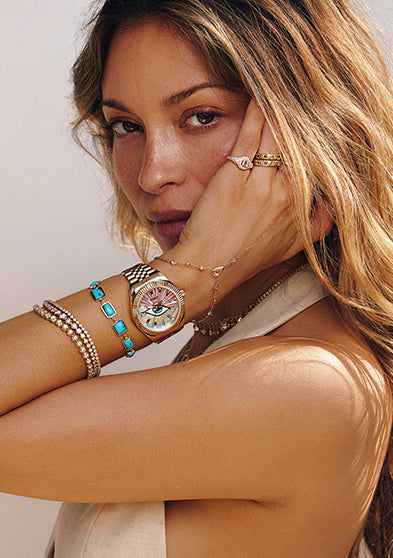 Woman wearing the Timex x Jacquie Aiche gold watch with a pink and multicolored mother-of-pearl dial featuring an eye motif, styled with layered gold and turquoise bracelets, posing with her hand near her face against a soft neutral background.