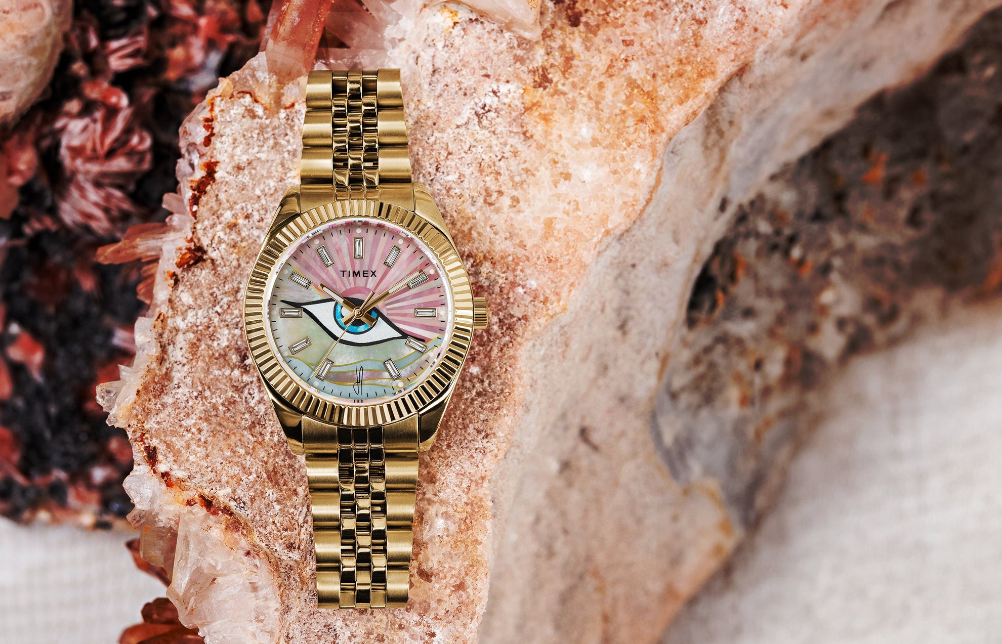 Timex x Jacquie Aiche collaboration watch with a gold bracelet and bezel, featuring a pink and multicolored mother-of-pearl dial with a central eye motif, displayed on a textured crystal and stone background.