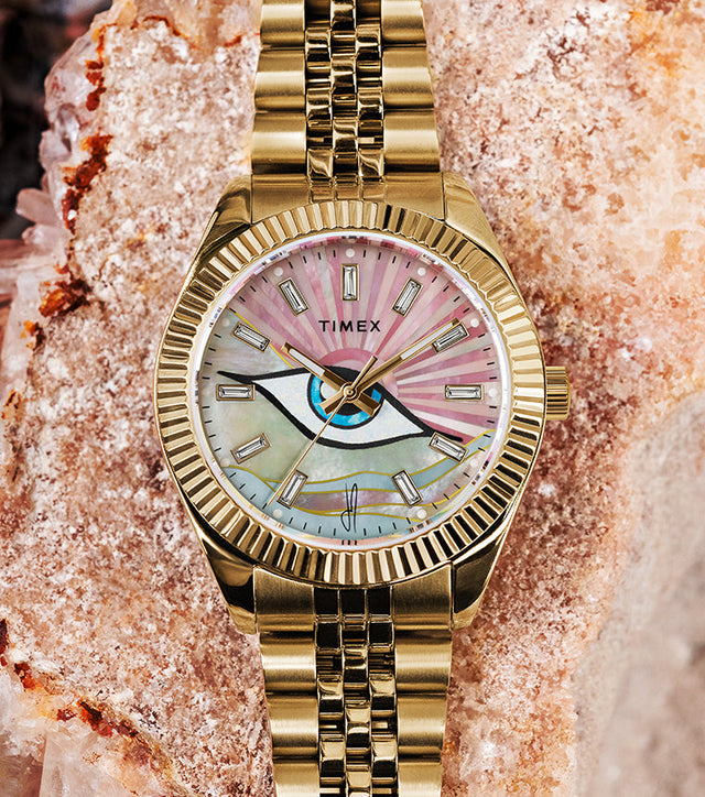 Timex x Jacquie Aiche collaboration watch with a gold bracelet and bezel, featuring a pink and multicolored mother-of-pearl dial with a central eye motif, displayed on a textured crystal and stone background.
