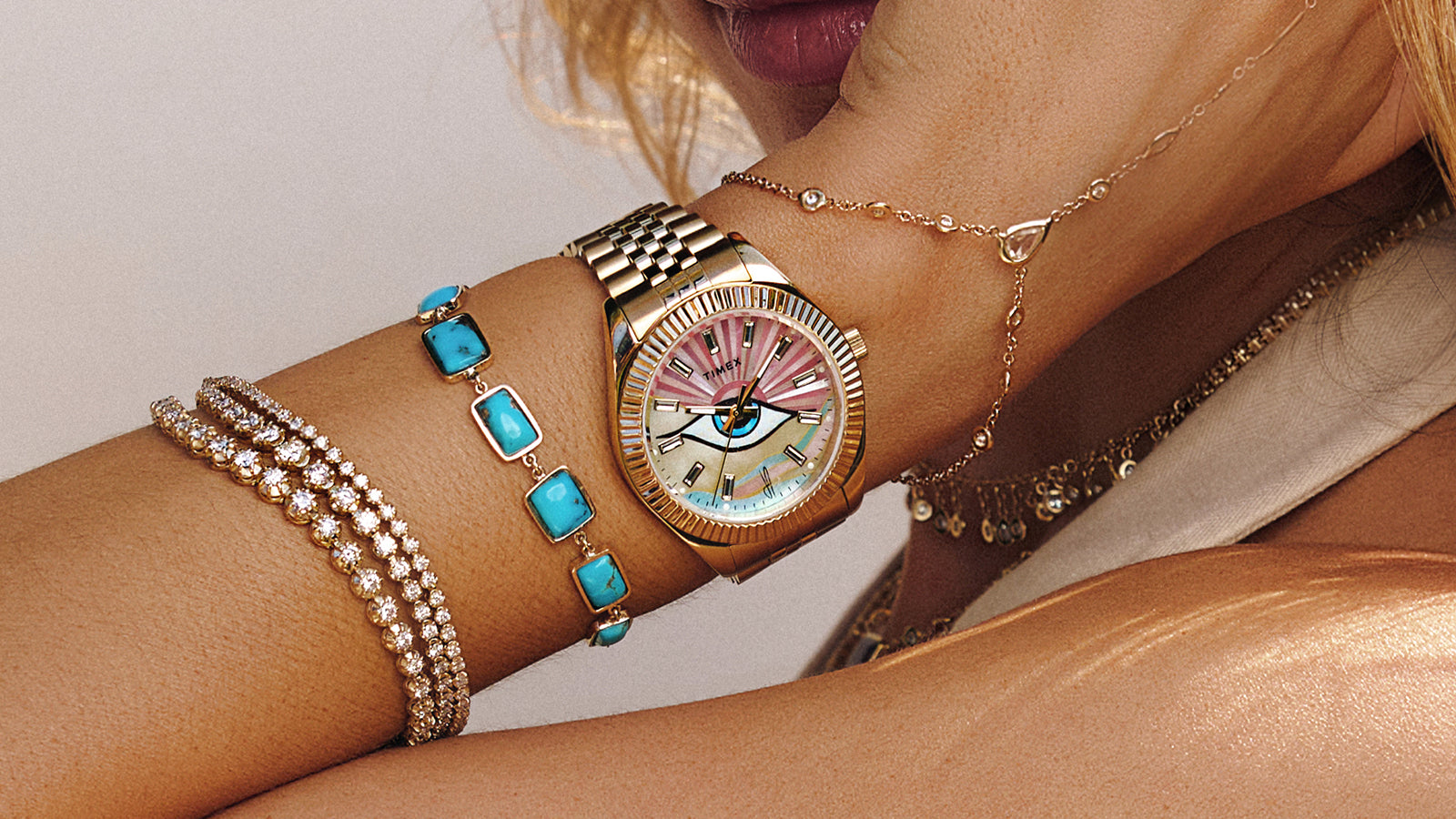 Woman wearing the Timex x Jacquie Aiche gold watch with an eye motif dial, styled with layered turquoise and diamond bracelets, posing with her wrist raised near her face against a neutral background.