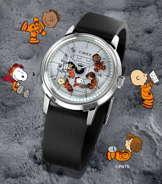 A Timex x Peanuts® Marlin® Automatic watch placed on a textured moon-like surface. The watch dial features Snoopy, Charlie Brown, Lucy, Franklin, and Peppermint Patty in orange space suits floating inside a space capsule. Additional Peanuts characters float around the watch on the lunar surface background.