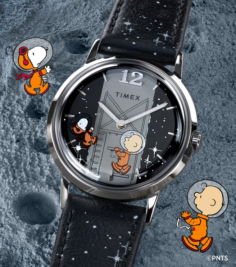 A Timex x Peanuts® Marlin® Hand-Wound watch displayed on a textured moon surface background. The black dial shows Charlie Brown climbing a space module ladder and Snoopy in a space helmet floating beside him. The watch has a black leather strap decorated with white stars.
