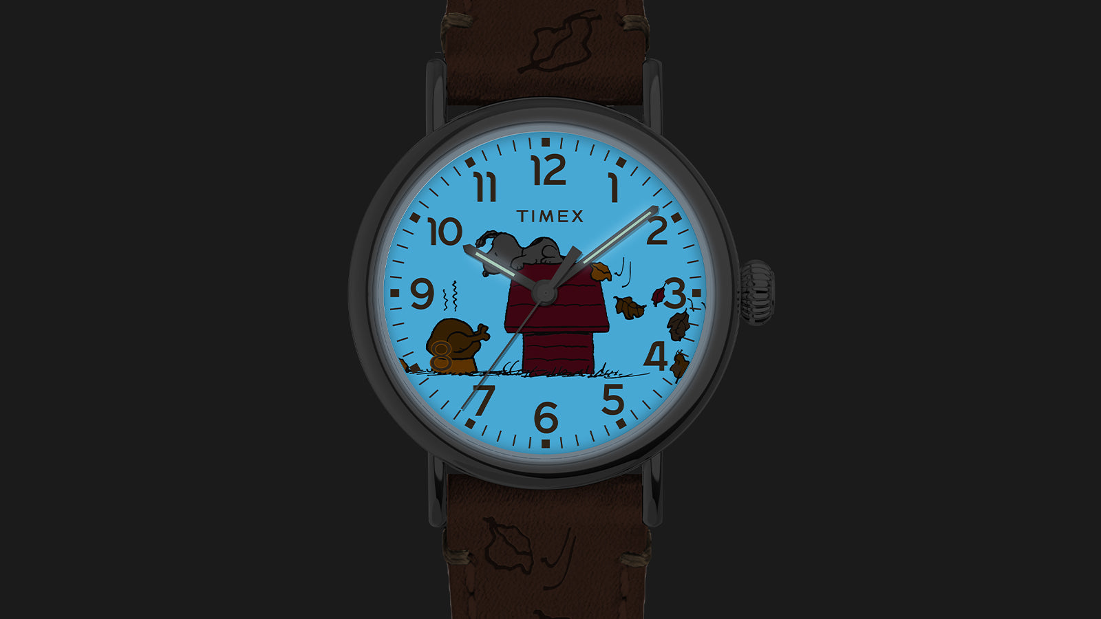 The Timex x Peanuts® Standard Thanksgiving watch illuminated with Timex INDIGLO® light, glowing blue in the dark to reveal Snoopy, Woodstock, and fall leaves in detail.