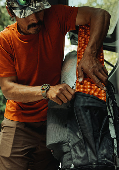 A man in an orange shirt and cap wears the TIMEX® IRONMAN® Flix x Huckberry while outdoors, packing camping gear. The rugged watch is shown as part of his active outdoor lifestyle.