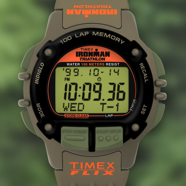 A detailed studio image of the TIMEX® IRONMAN® Flix x Huckberry watch. The screen shows bold black digits with an orange TIMEX FLIX logo on the strap. Features include a 100-lap memory, stopwatch, and water resistance to 100 meters.
