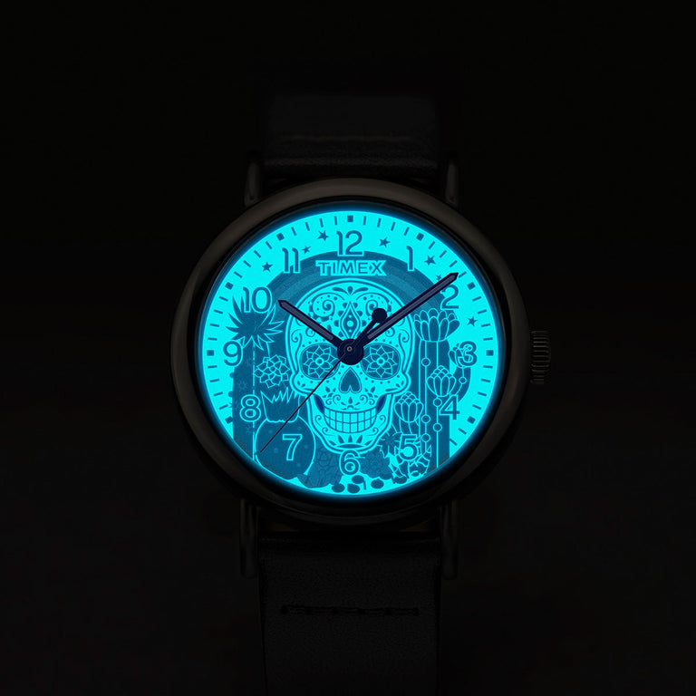 Timex Weekender Day of the Dead watch dial glowing in the dark with INDIGLO® backlight, revealing a bright blue sugar skull and floral details.