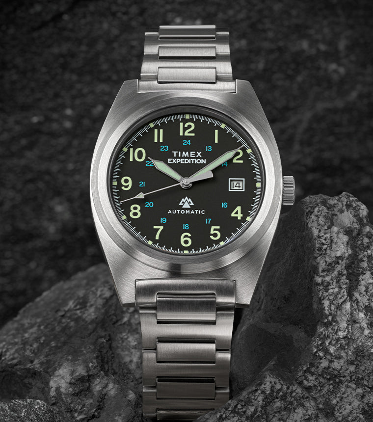 A Timex Expedition® Capstone Automatic watch with a brushed stainless-steel case and bracelet rests among rugged gray rocks. The black dial features luminous green Arabic numerals, a date window at 3 o’clock, and bold hands designed for outdoor durability.