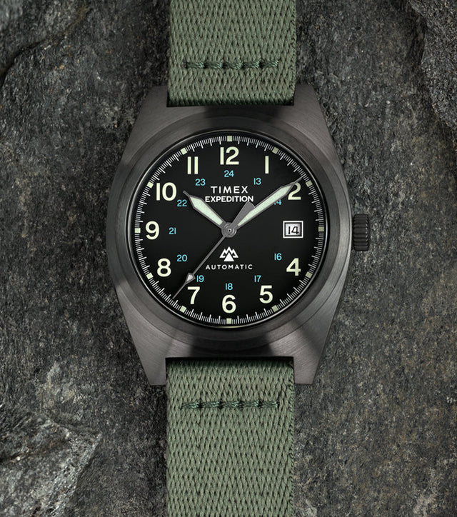 A Timex Expedition® Capstone Automatic watch with a matte gunmetal case and green fabric strap rests among rugged stone surfaces. The black dial features luminous green numerals, blue 24-hour markings, and a date window at 3 o’clock, emphasizing its field-ready durability.
