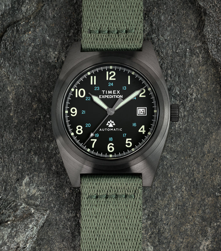 A Timex Expedition® Capstone Automatic watch with a matte gunmetal case and green fabric strap rests among rugged stone surfaces. The black dial features luminous green numerals, blue 24-hour markings, and a date window at 3 o’clock, emphasizing its field-ready durability.